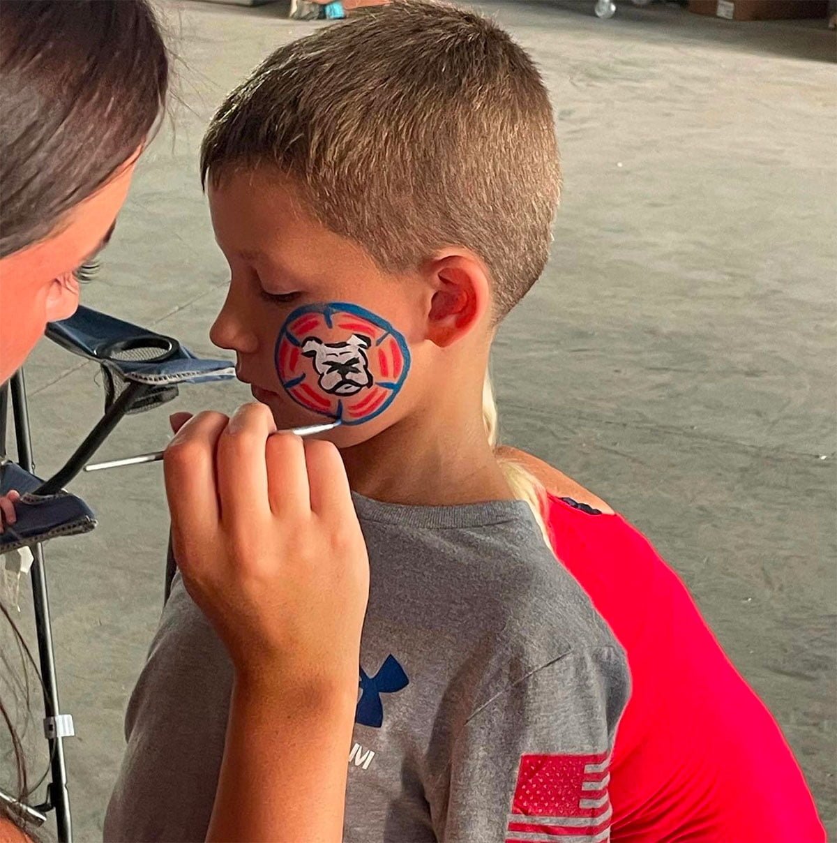 A young boy getting his face painted of a bulldog logo by a woman.